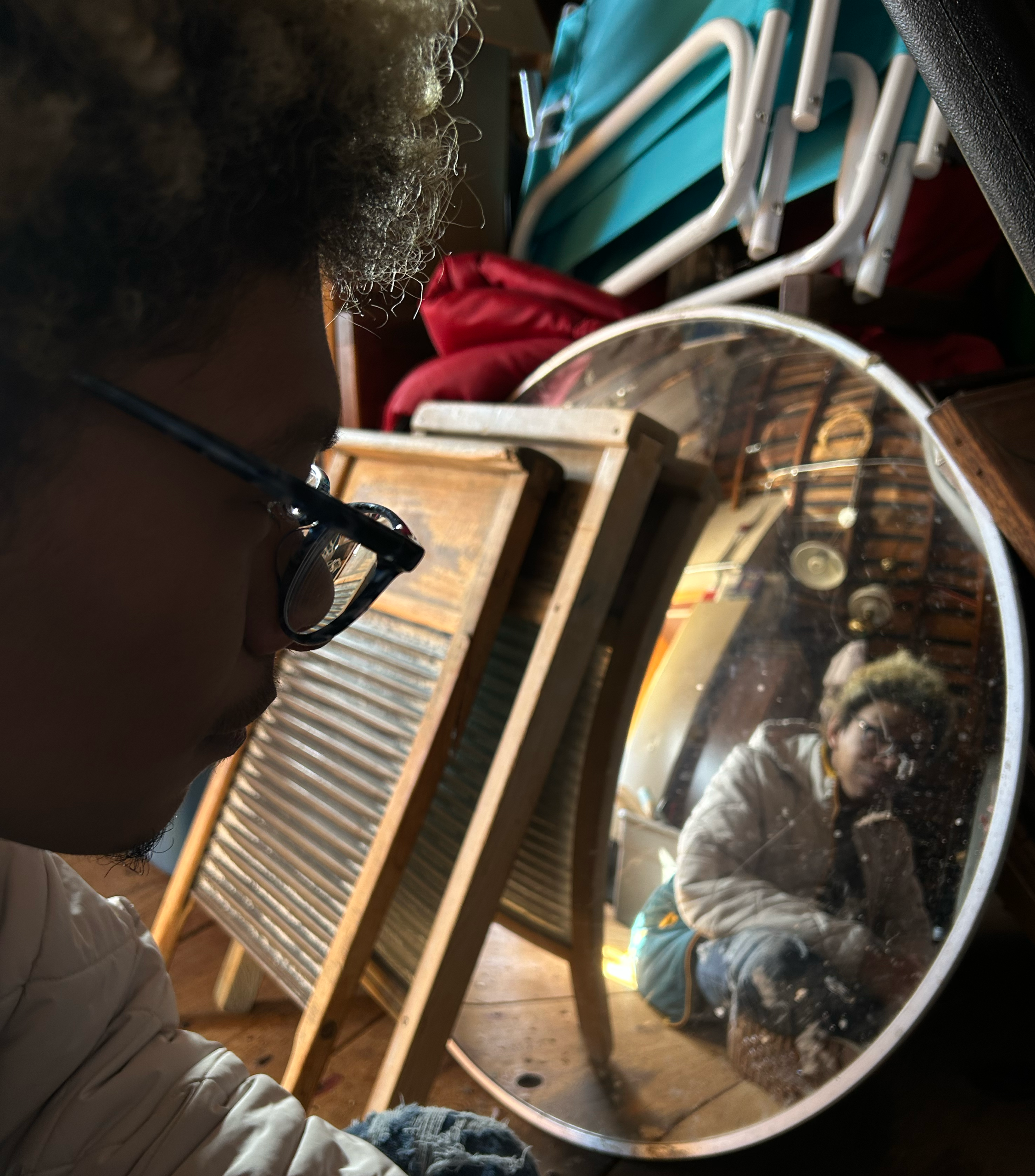 A person sitting on the ground while looking off to the side. The photographer is focusing on the mirror more so than the person. There is clutter in the background.